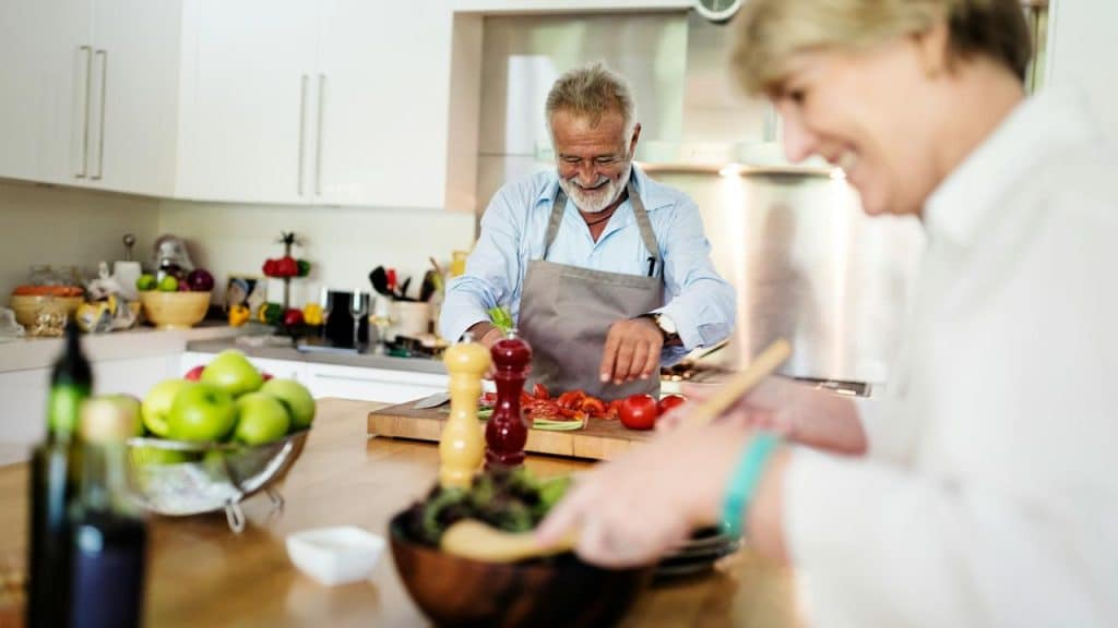 A smiling elderly man chopping tomatoes while a woman mixes salad in a bright kitchen.