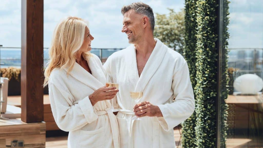A smiling couple in white bathrobes holding wine glasses on a terrace.