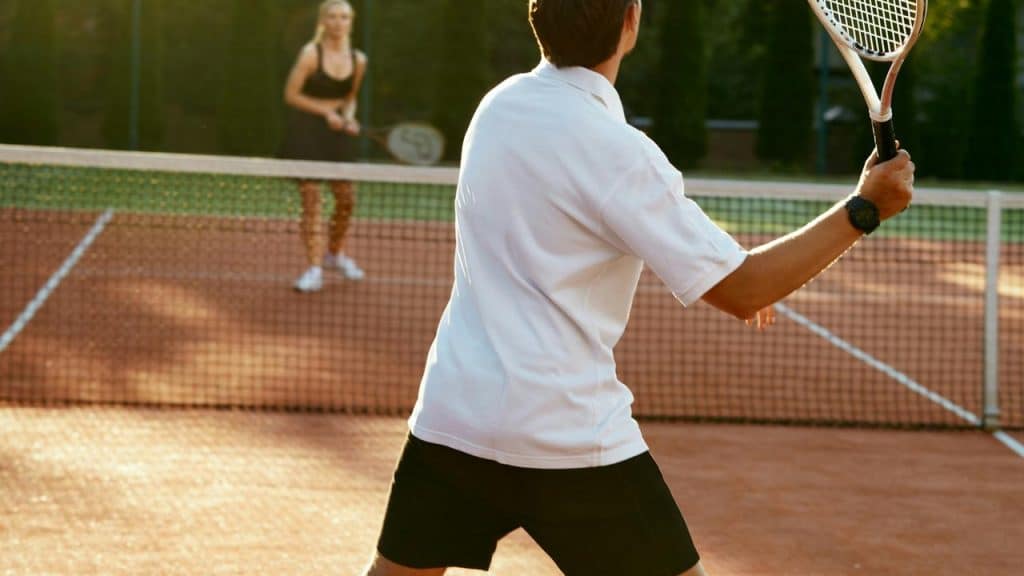 A man playing tennis on an outdoor court with a woman opponent in the background.