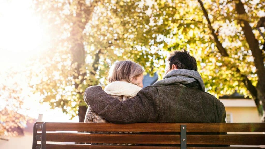 A couple sitting on a bench outdoors with the manโs arm around the woman.