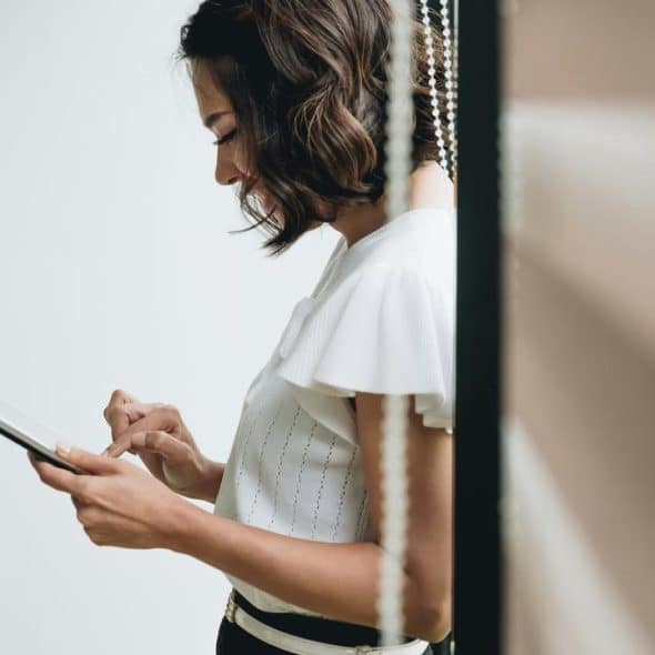 A woman using a smartphone indoors while her back rests on the window blinds.
