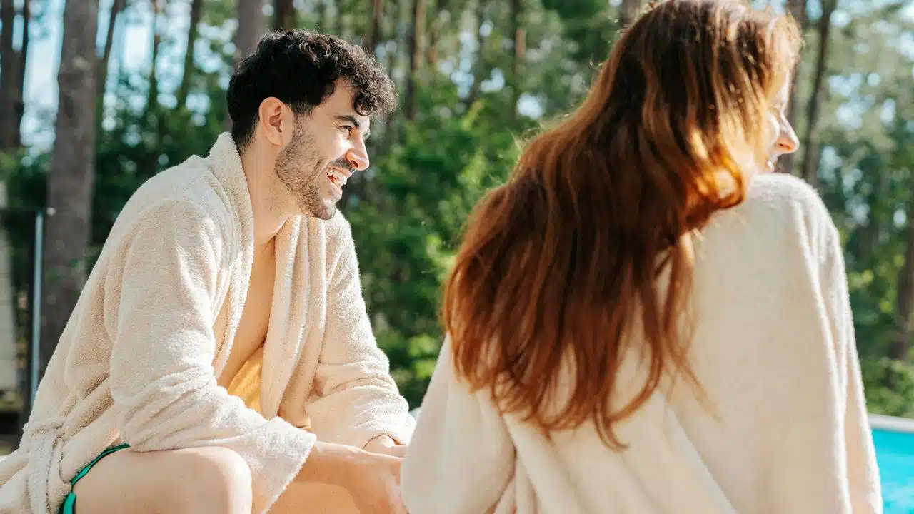 A man and woman in bathrobes talking by a pool.