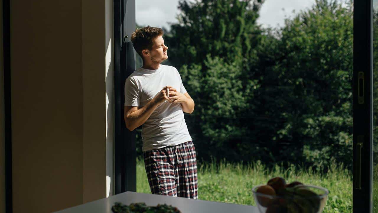 A man in pajamas holding a cup and looking outside through a large doorway..