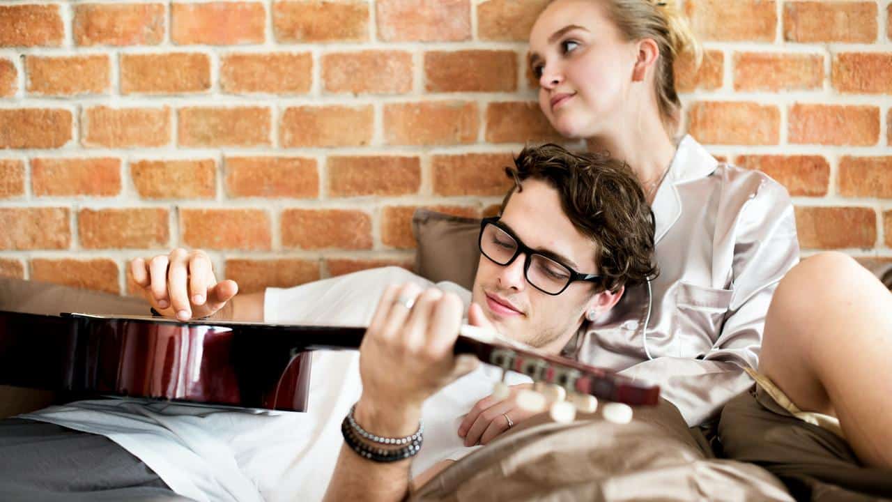A couple relaxing on a bed while the man plays guitar.