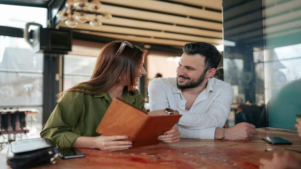 A man and a woman sitting at a table in a cafe, looking at a menu together and smiling at each other.