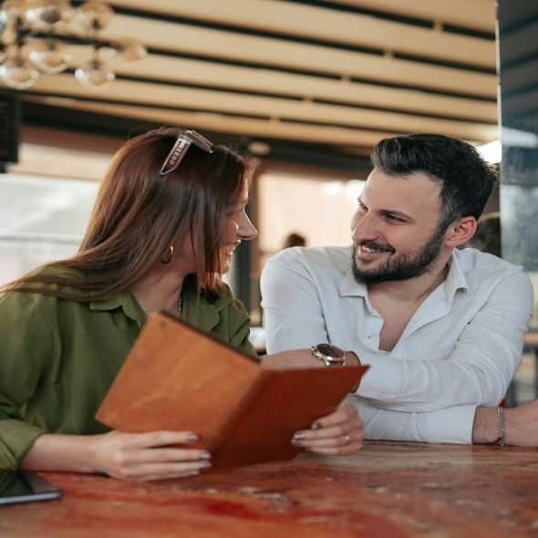 A man and a woman sitting at a table in a cafe, looking at a menu together and smiling at each other.