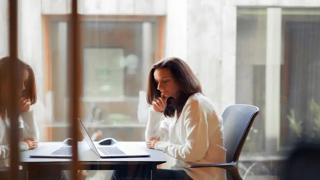 A woman sitting at a desk in a modern office, looking at her laptop with a thoughtful expression.