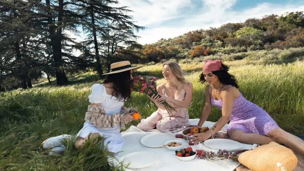 Three women having a picnic in a grassy field, with one holding a bouquet of flowers.