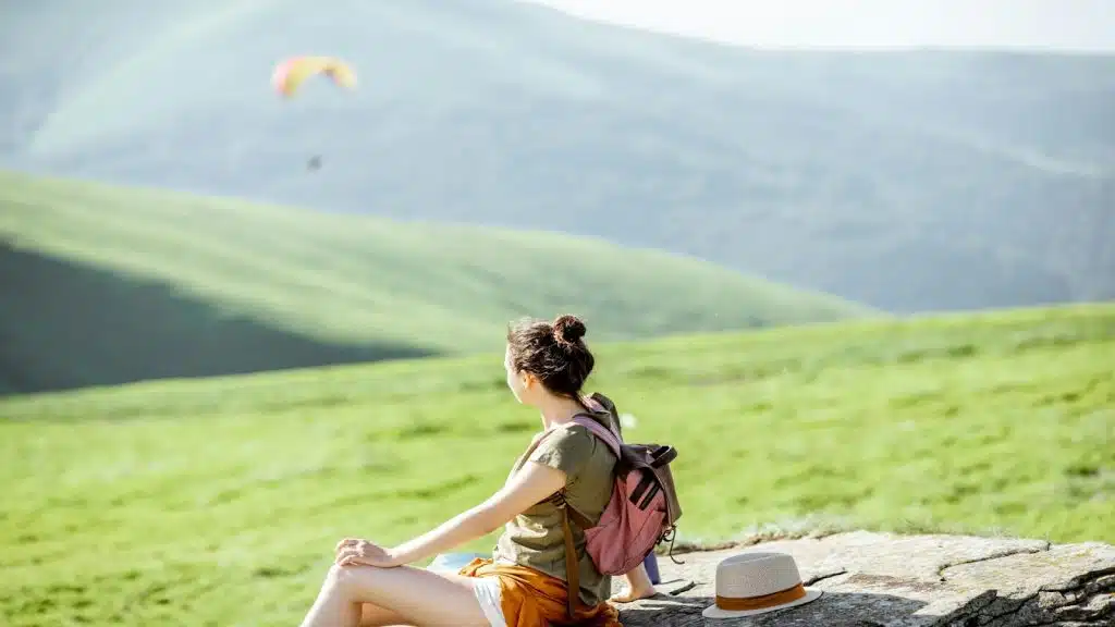 A woman with a backpack and a hat standing on a mountain ridge, looking out at a scenic view.