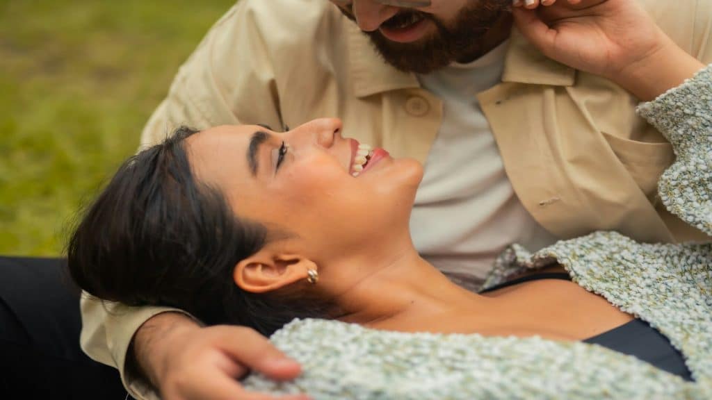 A  man and a woman relaxing in the grass, with the woman's head resting on the man's stomach as they smile at each other.
