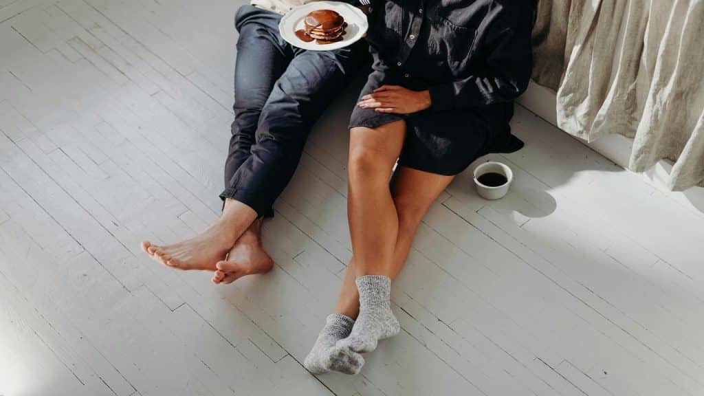 A man and a woman sitting on the floor, holding cups and a plate of pancakes with chocolate on them