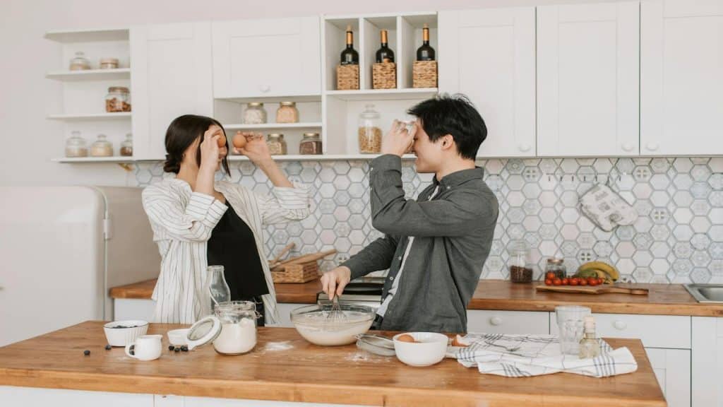 A man and a woman in a kitchen, holding eggs and smiling at each other.
