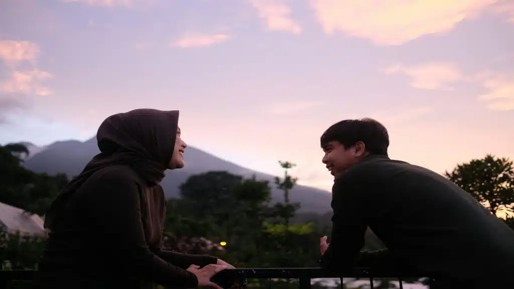 A man and a woman sitting on a rock with a sunset in the background, talking and smiling at each other.