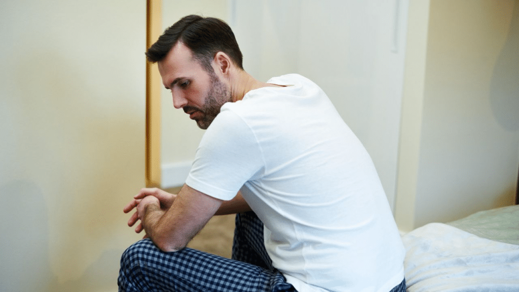 A man in a white t-shirt and plaid pajama pants sits on a bed with a sad and thoughtful expression.