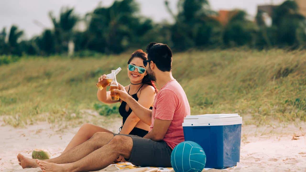 A couple sitting on the beach clinking beer bottles.