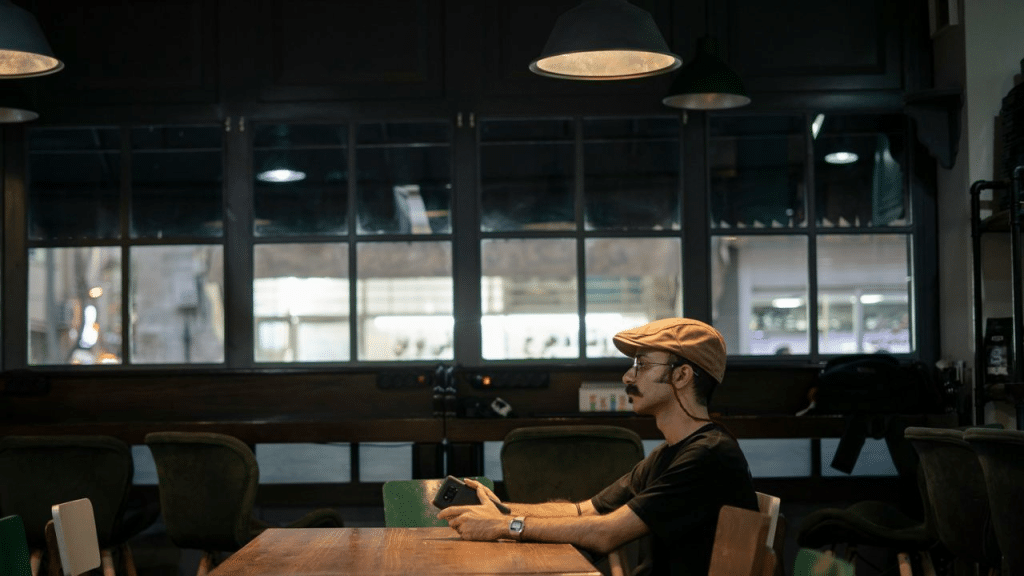 A man wearing a flat cap sits at a dark wooden table in a cafe, looking at his phone.