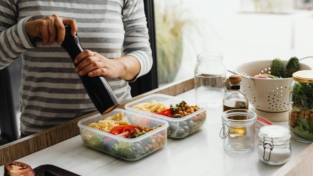 A person preparing pasta salad meals in containers with vegetables and chickpeas.