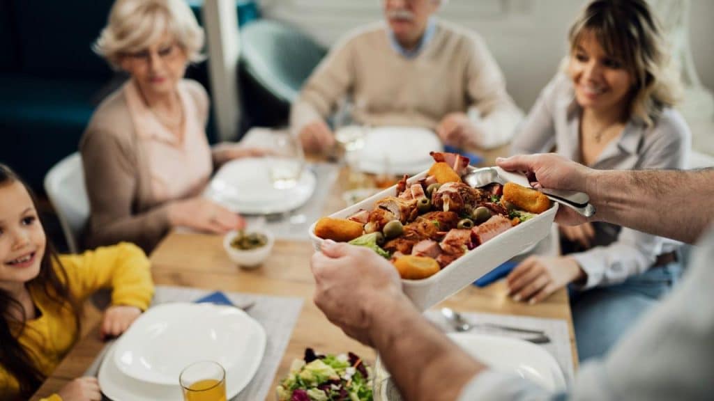 A family sitting at a table while someone serves a dish of food.