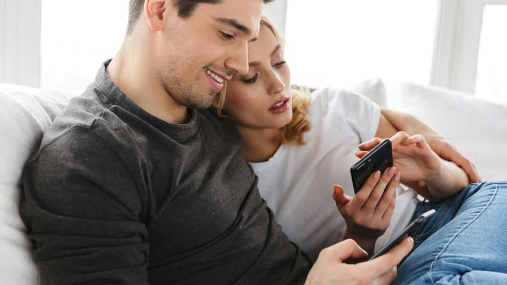 A couple sitting together on a couch, looking at a phone and smiling.