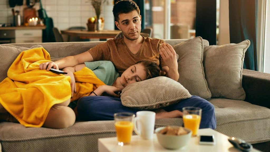 A couple relaxing on a couch with one person sleeping under a yellow blanket.