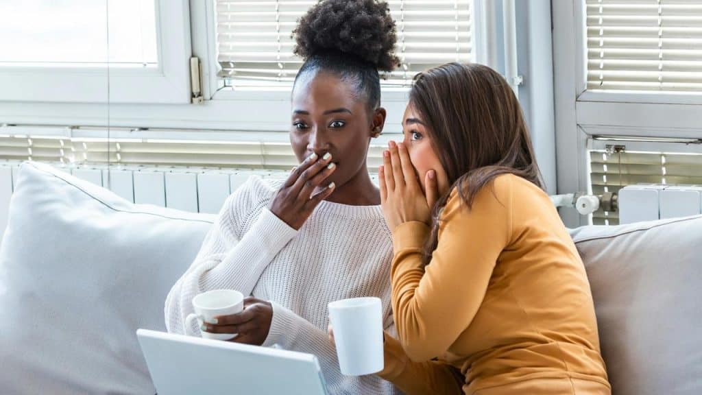 A woman whispering to another woman, who looks surprised.