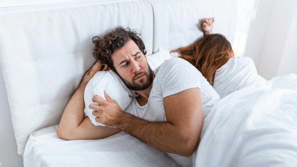 A troubled man lying awake in bed, his partner turned away from him.