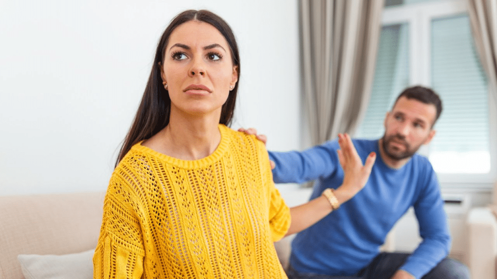 A worried woman in a yellow sweater, with a man reaching for her from behind.