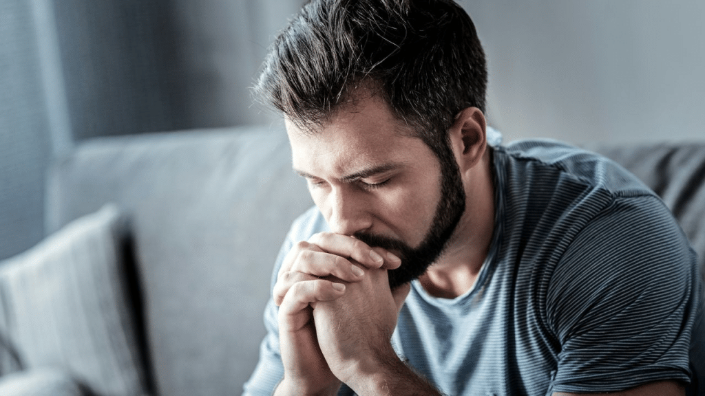 A pensive, bearded man sitting alone on a sofa, looking down sadly.