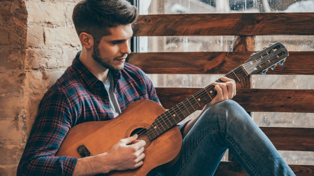 A focused man with a beard sits and plays an acoustic guitar by a window.
