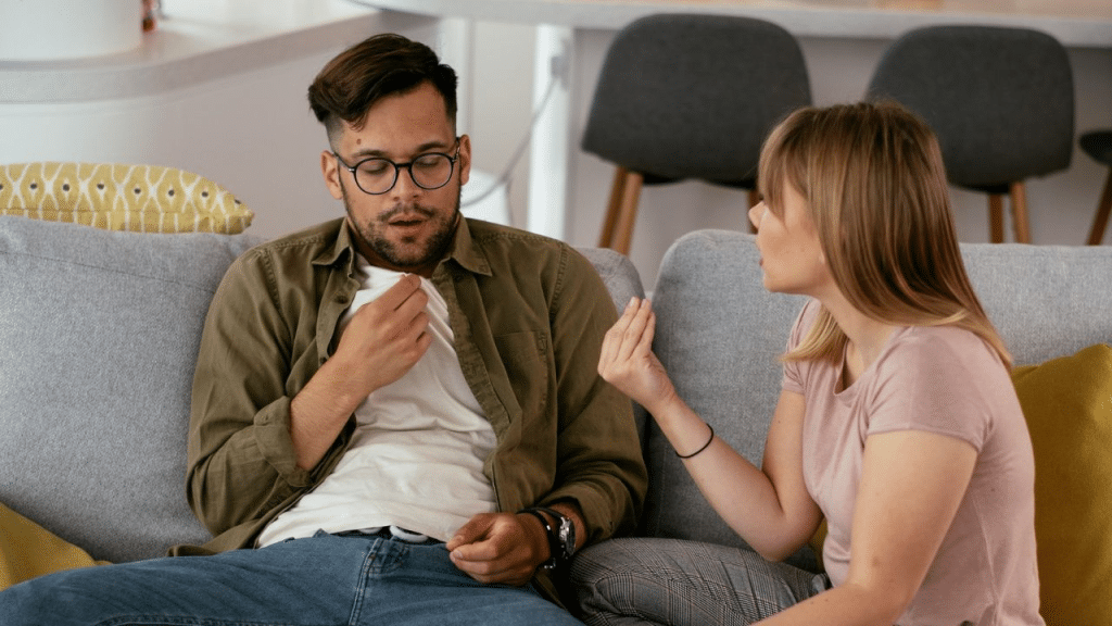 A tired man on a sofa holds a tissue while a woman talks to him.