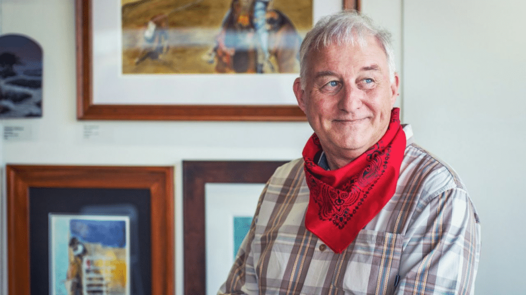 A handsome, smiling older man with a red bandana around his neck is looking to his left.