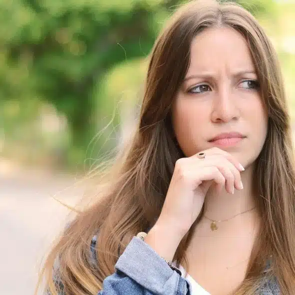 A young woman outdoors looking thoughtful with her hand on her chin.