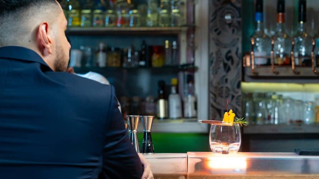 A man in a dark suit sitting at a bar with a cocktail in front of him.