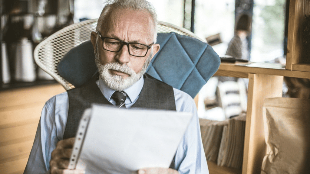 A thoughtful, senior Caucasian businessman with a beard and glasses is reading a document.