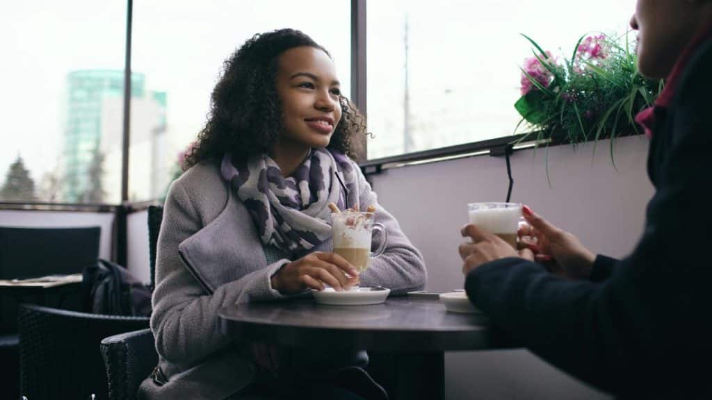 A woman in a coat and scarf smiling while having coffee at a café table.