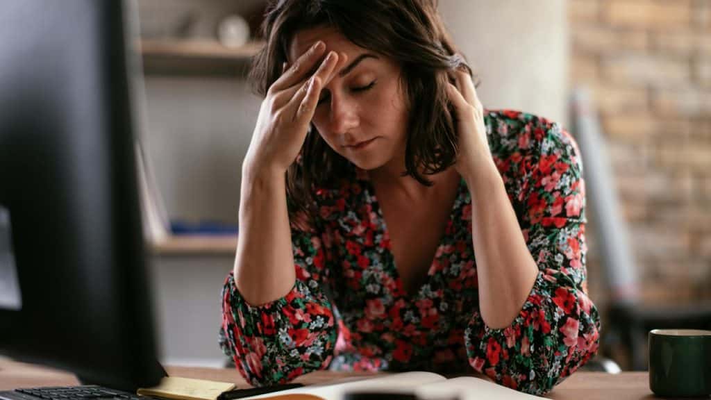 A woman in a floral dress sitting at a desk with her hand on her forehead.