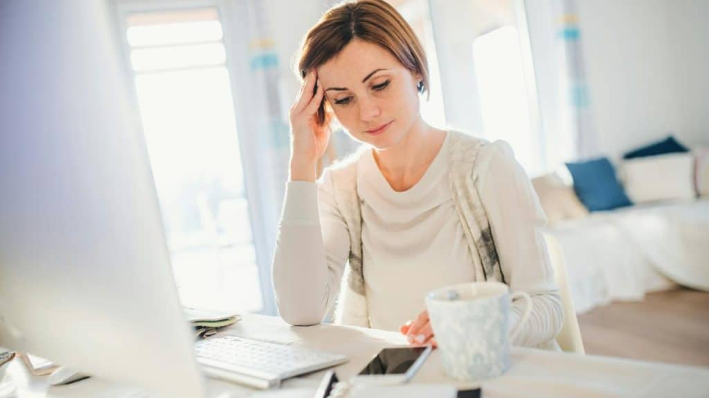 A woman in a white top sitting at a desk with her hand on her temple