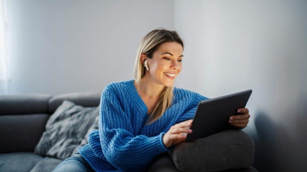 A woman in a blue sweater smiling while using a tablet with wireless earbuds in.