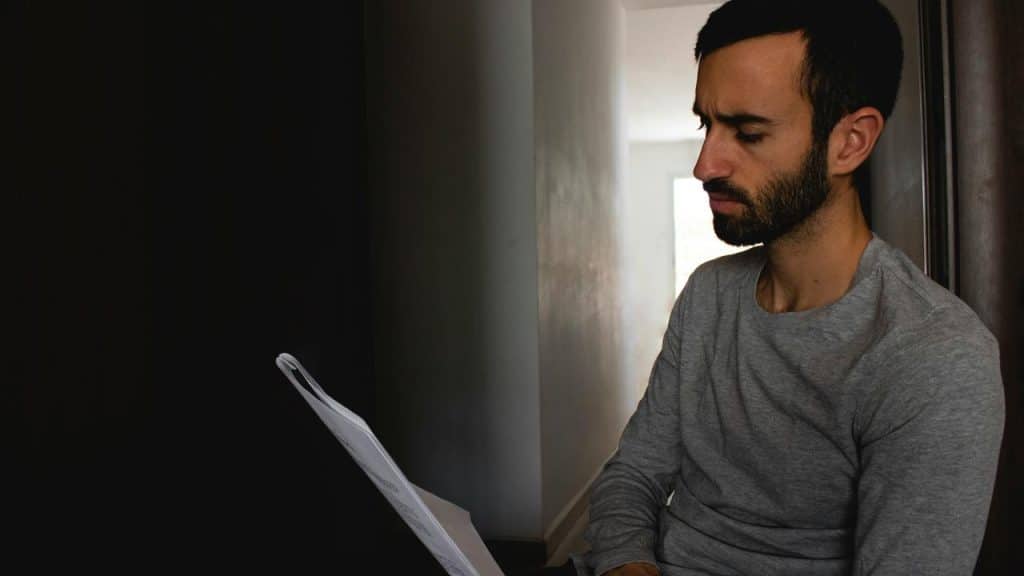 A man sitting indoors reading a document with a serious expression.