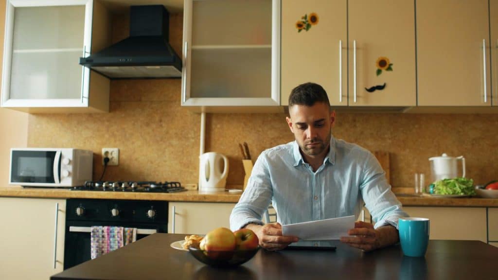 A man sitting in a kitchen reading a paper with a serious expression.