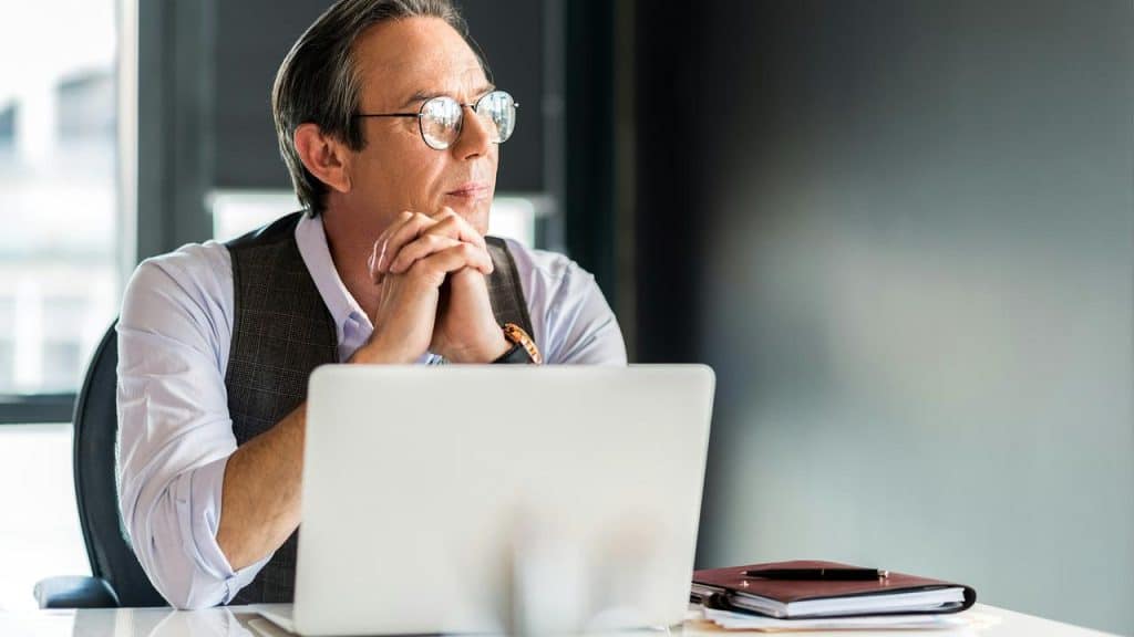 An older man wearing glasses sitting at a desk with a laptop, looking thoughtful.
