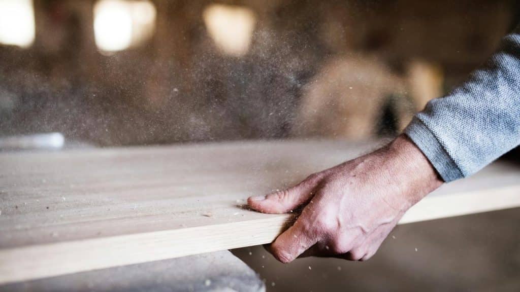 A close-up of a man’s hand working with a wooden board covered in sawdust.