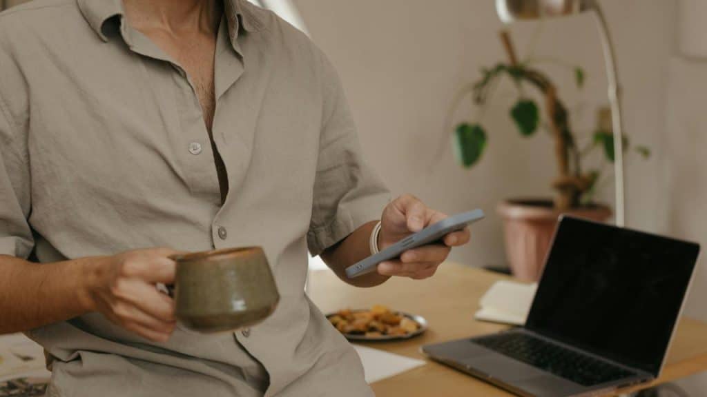 A man holding a mug and a phone while sitting at a desk with a laptop.