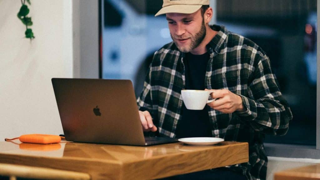 A man wearing a cap and flannel shirt working on a laptop while holding a cup of coffee.