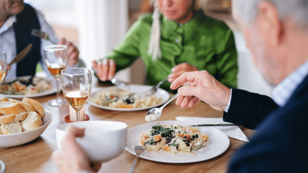 A diverse group of people is seated at a table, eating pasta and drinking wine.