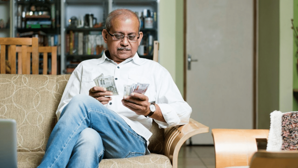 A man is seated on a sofa, looking at cash and holding Indian rupee notes in each hand.