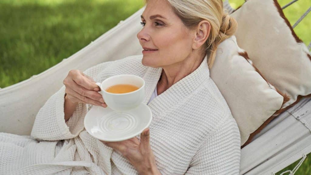 A woman in a white robe relaxing on a hammock while holding a cup of tea.