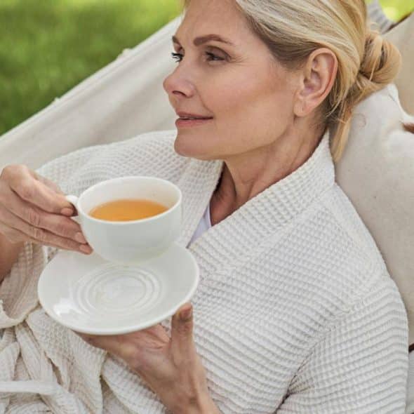 A woman in a white robe relaxing on a hammock while holding a cup of tea.