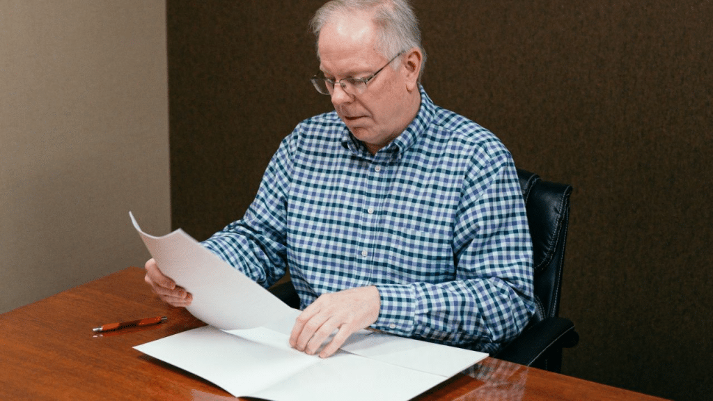 A thoughtful older man with glasses is seated at a wooden desk and looking at papers.