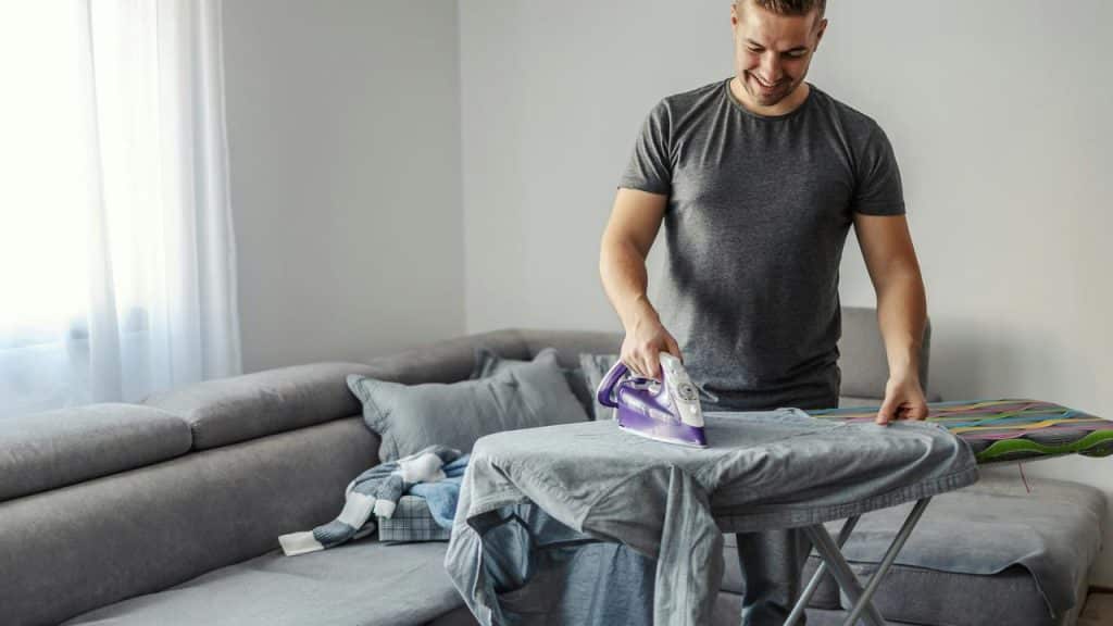 A man smiling while ironing clothes in a living room.